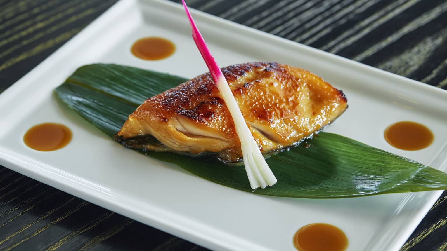 Black Cod Yuzu Miso fish fillet marinated, oven-baked on grilled seafood filet on leaf on white platter