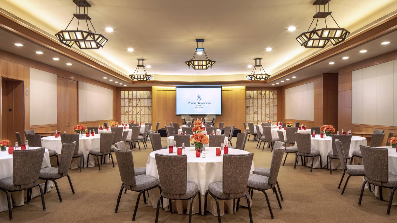 A large, wood panelled function room with circular tables set with a white tablecloth, red cups and orange flowers