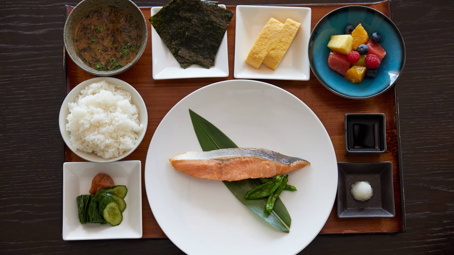 On wooden tray in dishes of various shapes and sizes, an alluring spread of food items, including fish, rice and fresh fruit.