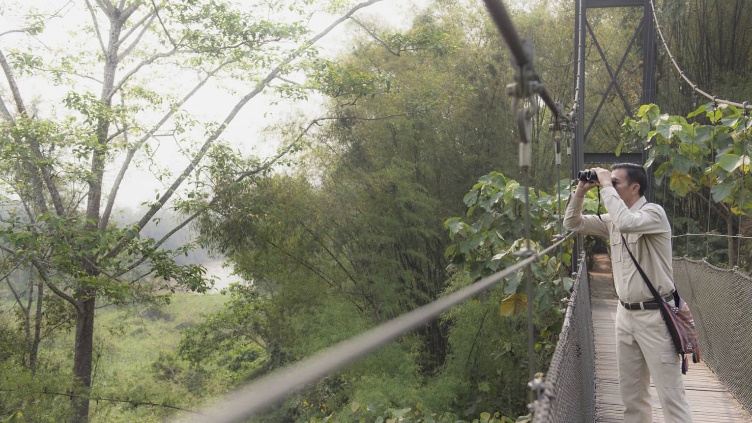 Man wearing artisan bag on suspension bridge surrounded by jungle looking through binoculars 