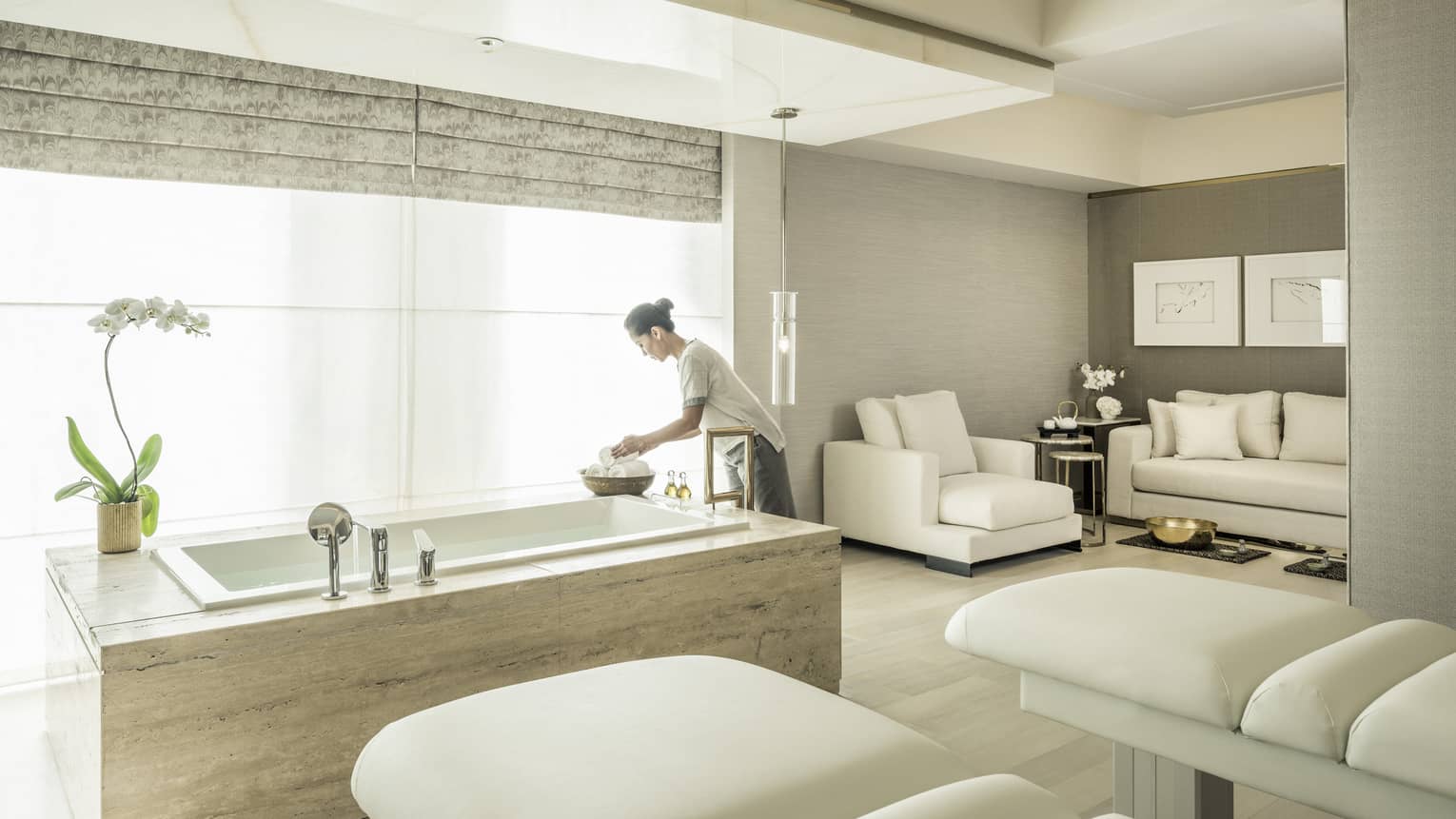 A spa attendant arranges some products in the spa suite, with two massage beds and a soaking tub in front