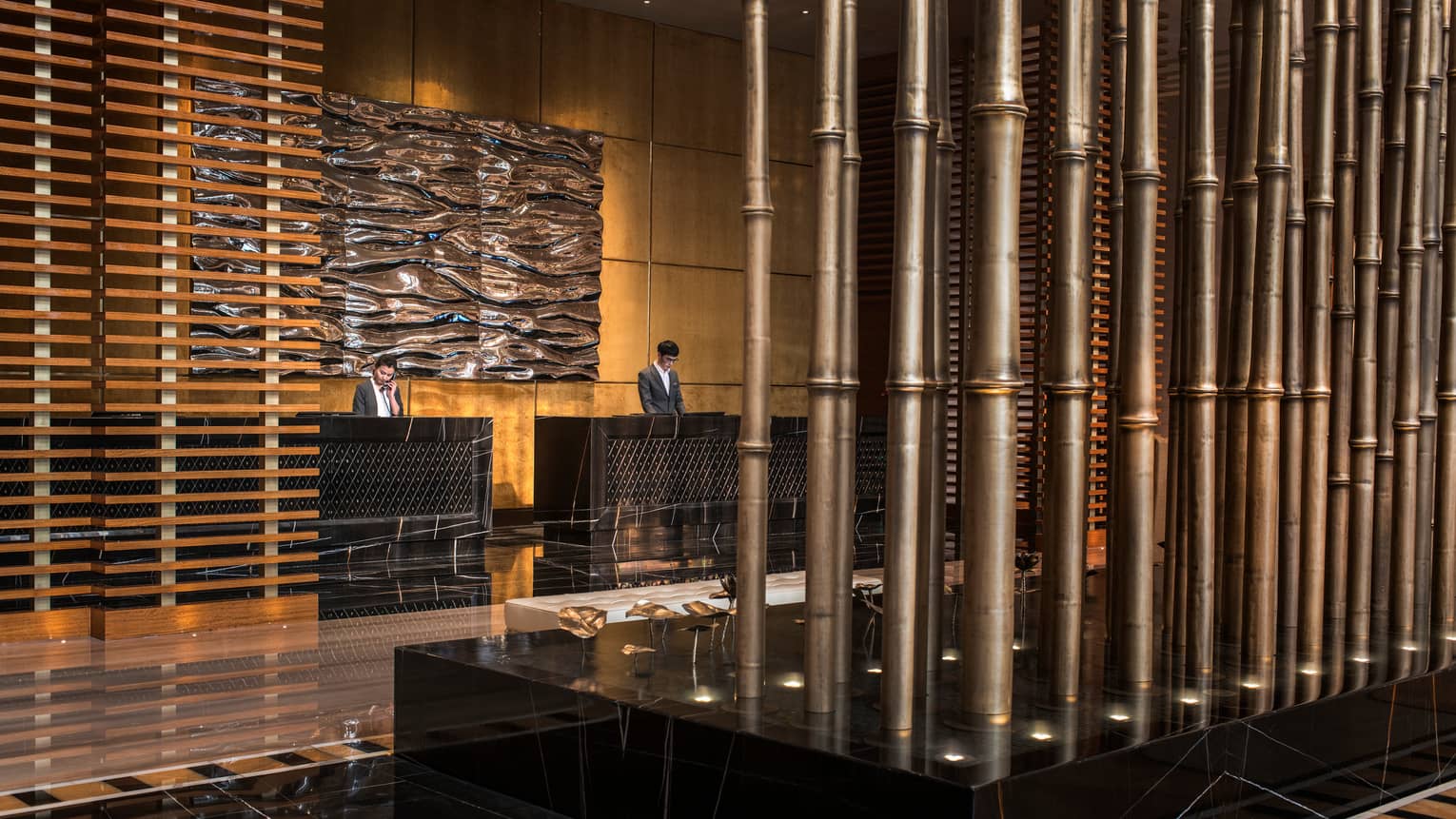 Two hotel staff behind reception desks under large wave-like wall sculpture, wood panel wall