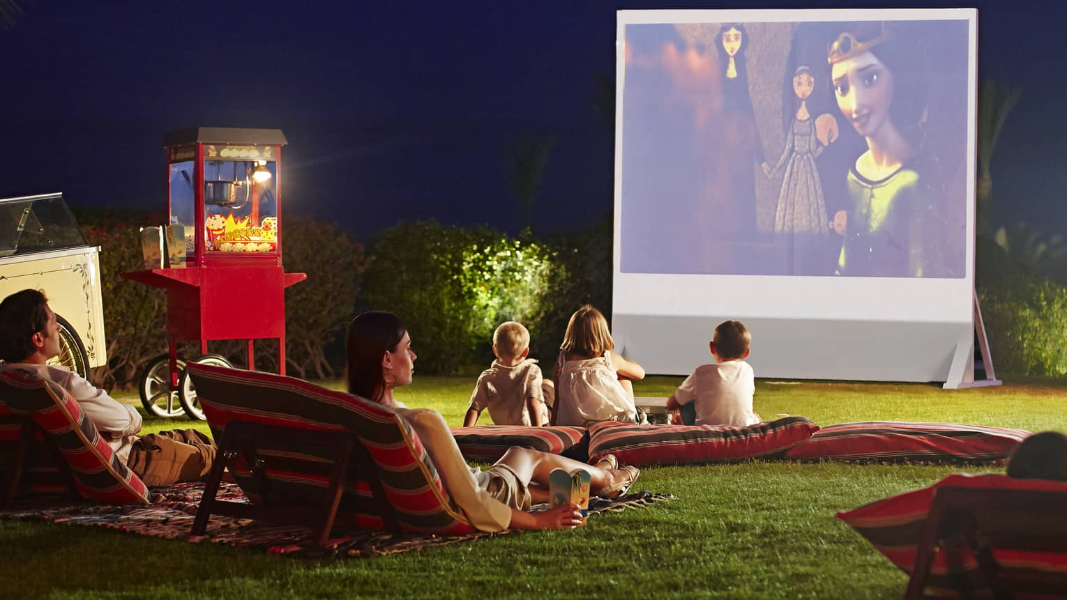 Children, parents lounge on pillows on grass in front of large outdoor movie screen at night