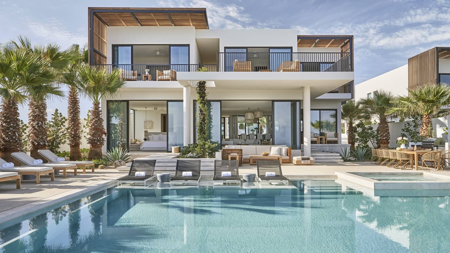 Modern luxury villa with a spacious pool area, surrounded by palm trees, featuring outdoor lounge chairs, a dining area, and floor-to-ceiling windows.