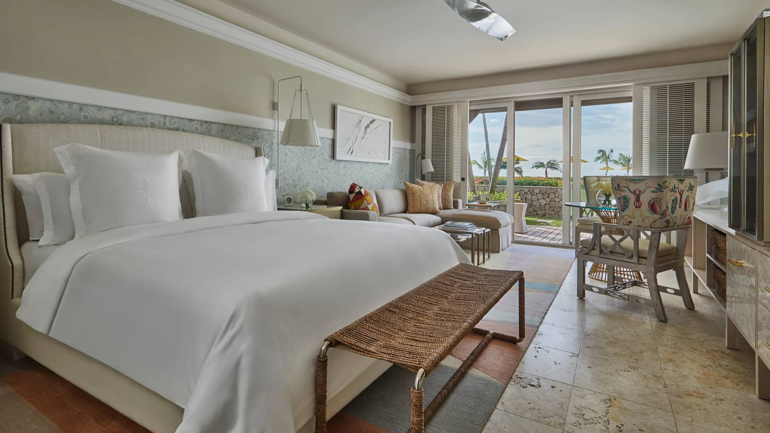 A room with a bed with white sheets, large window, small sofa and chair.