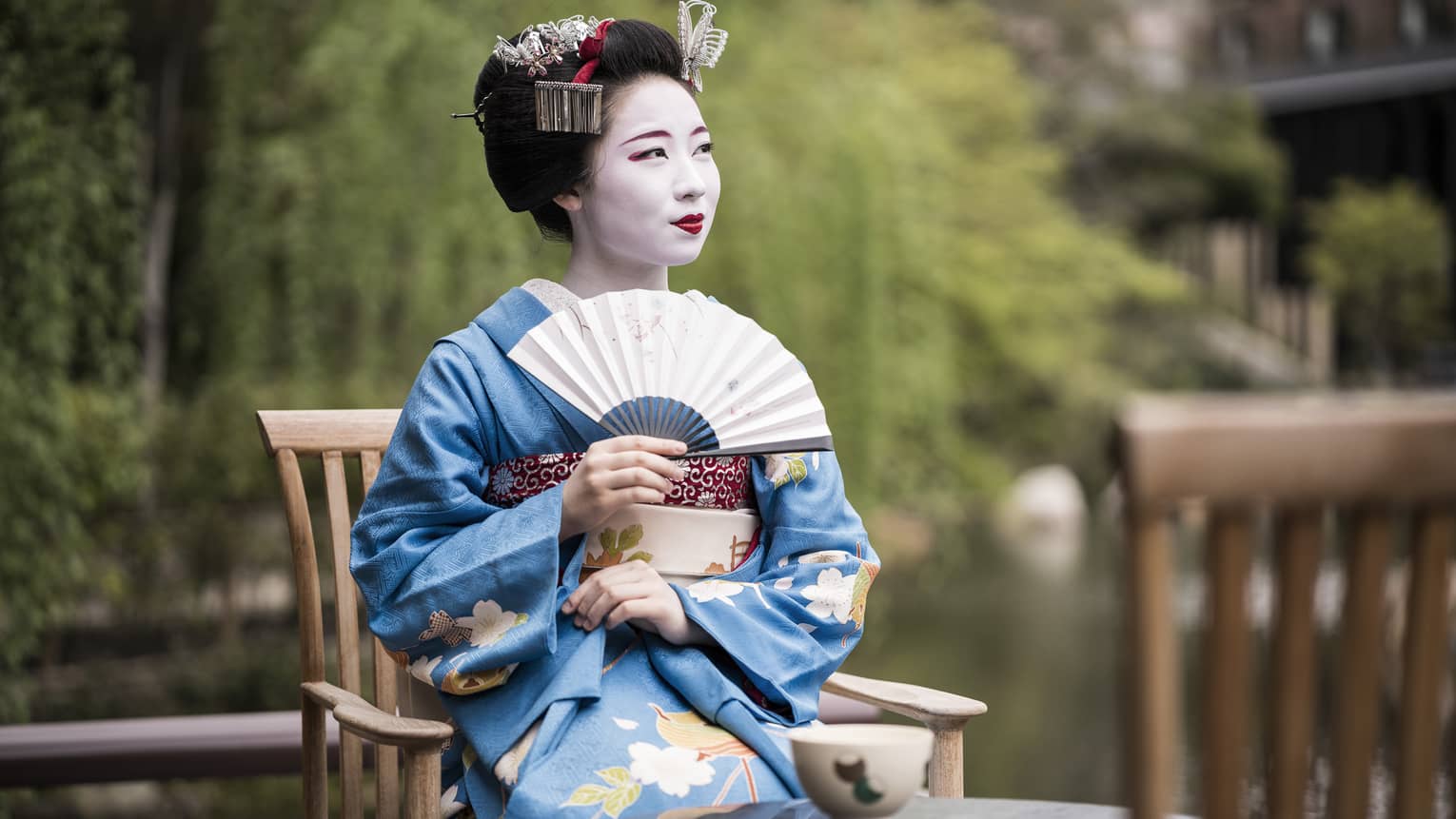 Geiko, a traditional Japanese entertainer, dressed in kimono and holding fan, sitting with cup of tea in garden