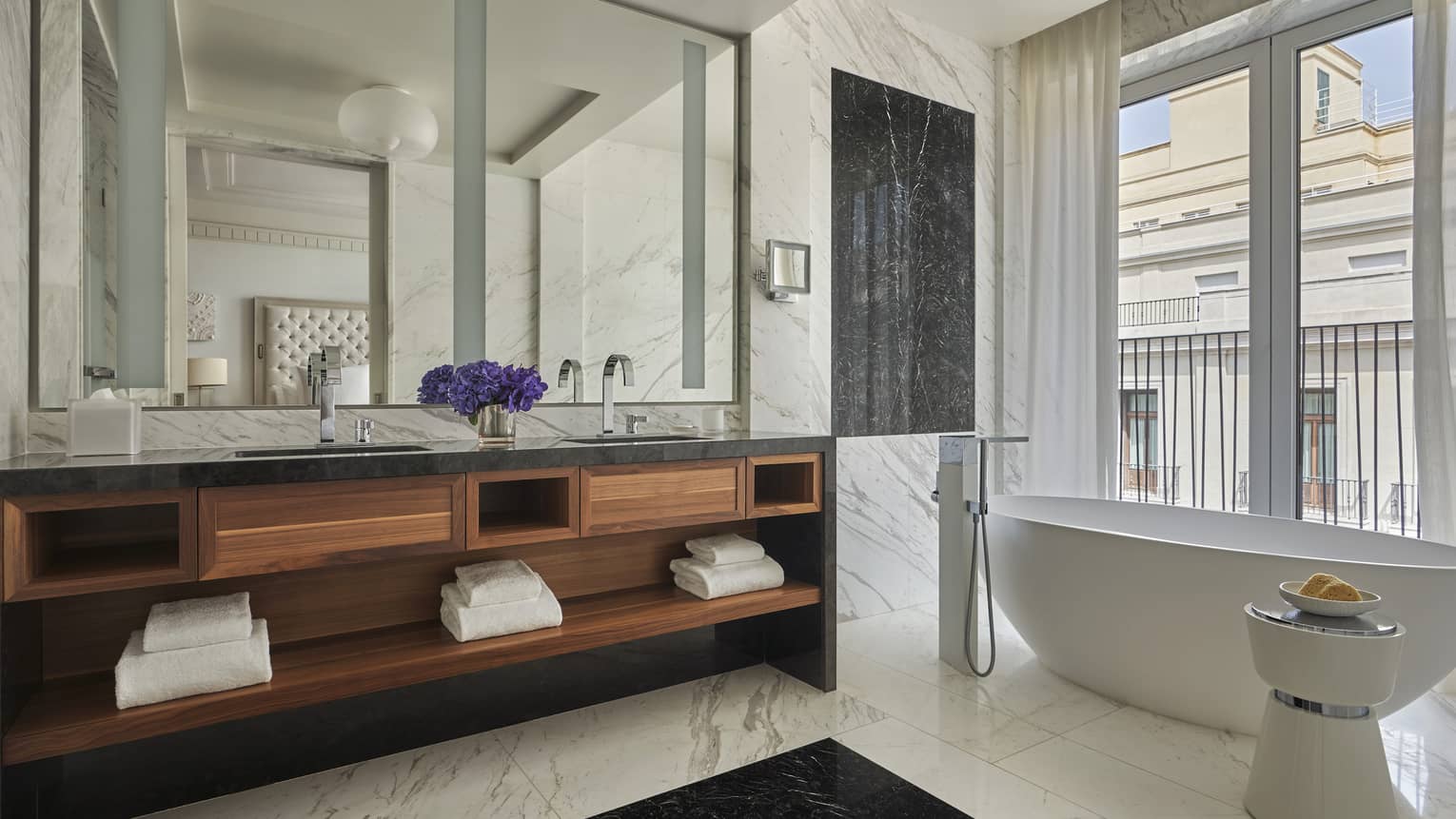 Bathroom with white marble walls and floors, oval white tub, wooden double vanity 