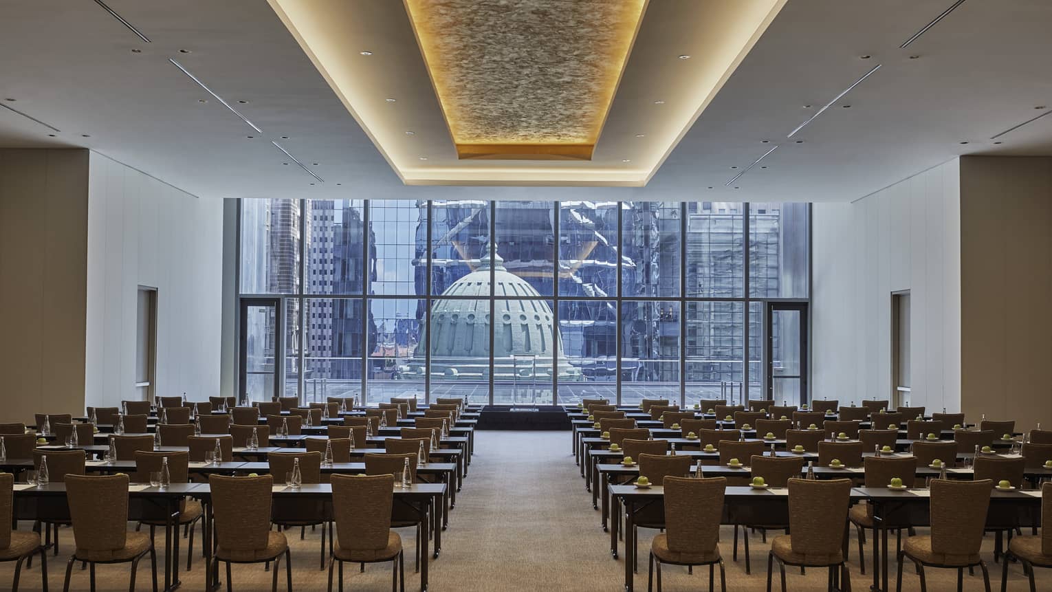 A large meeting room with rows of tables and chairs facing a podium with a wall of windows behind it.