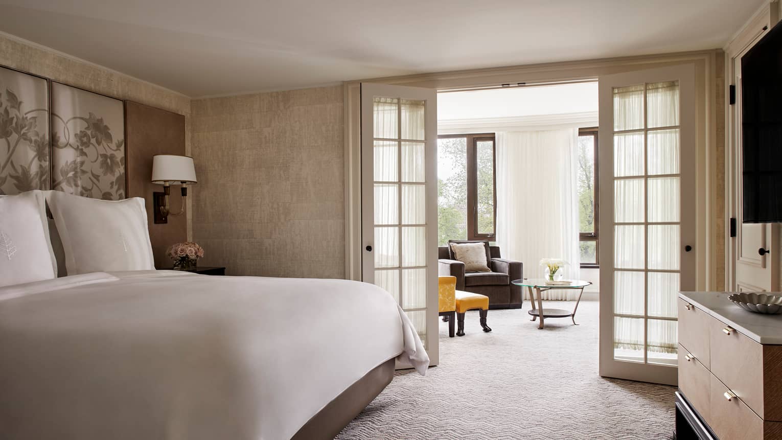 Senator Suite bed with white linens beside glass French doors leading to sunny living room with armchairs 
