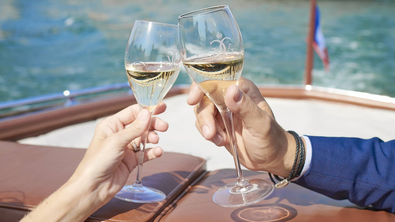 Two hands clink glasses of white wine at the back of a boat cruising on a clear-blue river near an ornate bridge.