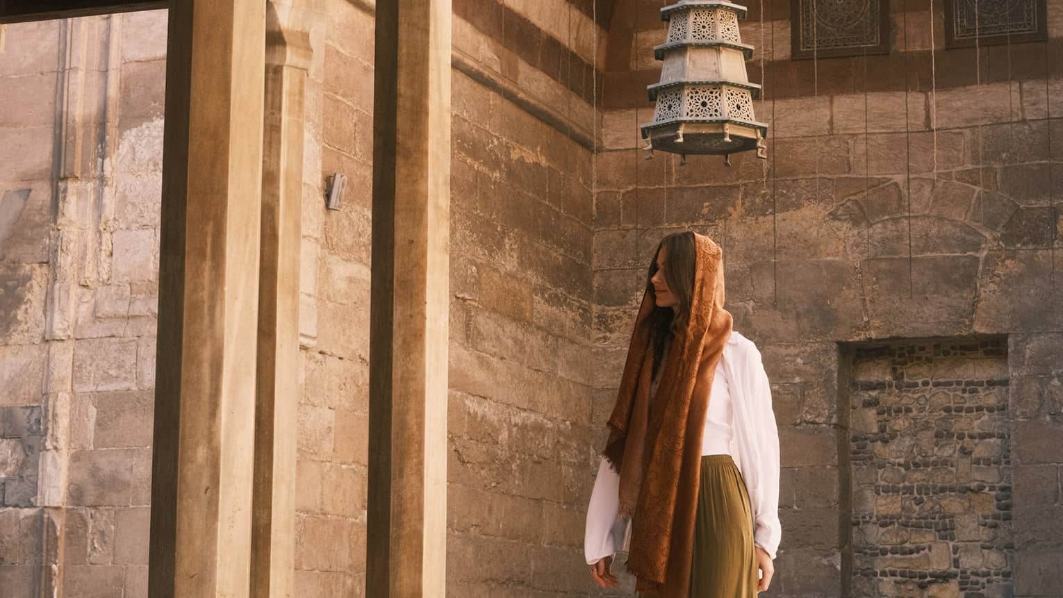 A person, head covered, stands under a pyramidal mosque lantern amid ornately carved walls and columns, a black cat nearby.