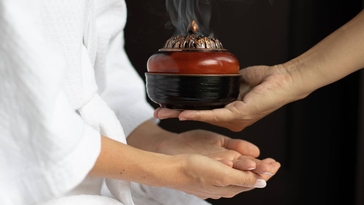 A person in a white robe receiving a steaming incense bowl from a therapist, creating a calming ambiance during a spa treatment.