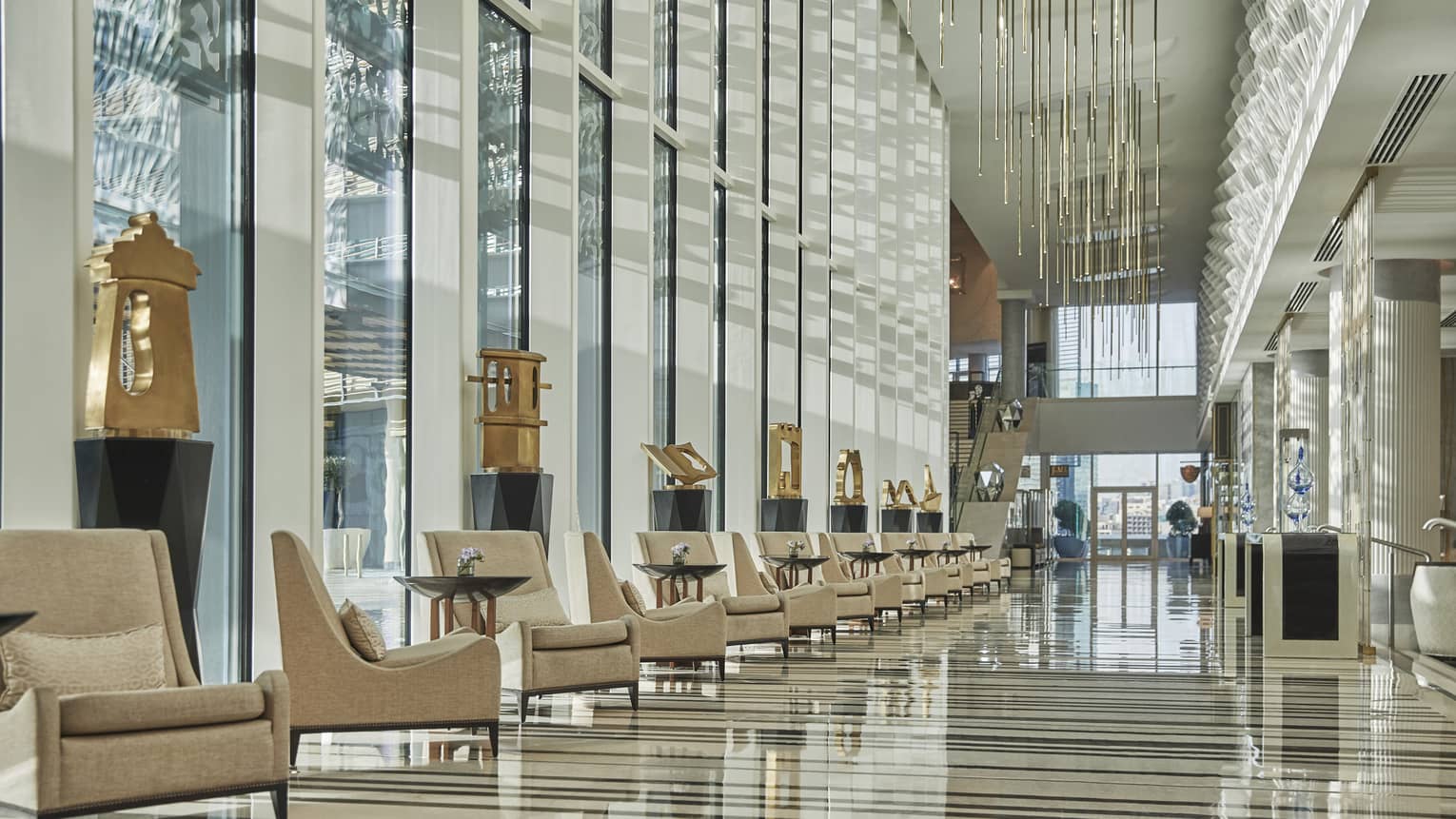 Hotel lobby with row of tan arm chairs, gold sculptures and floor-to-ceiling windows for natural light