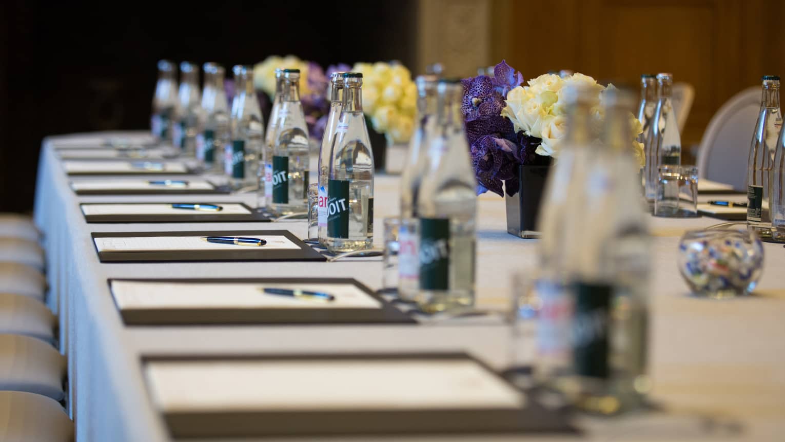 Row of meeting agendas, glass bottles, small vases with flowers on boardroom table
