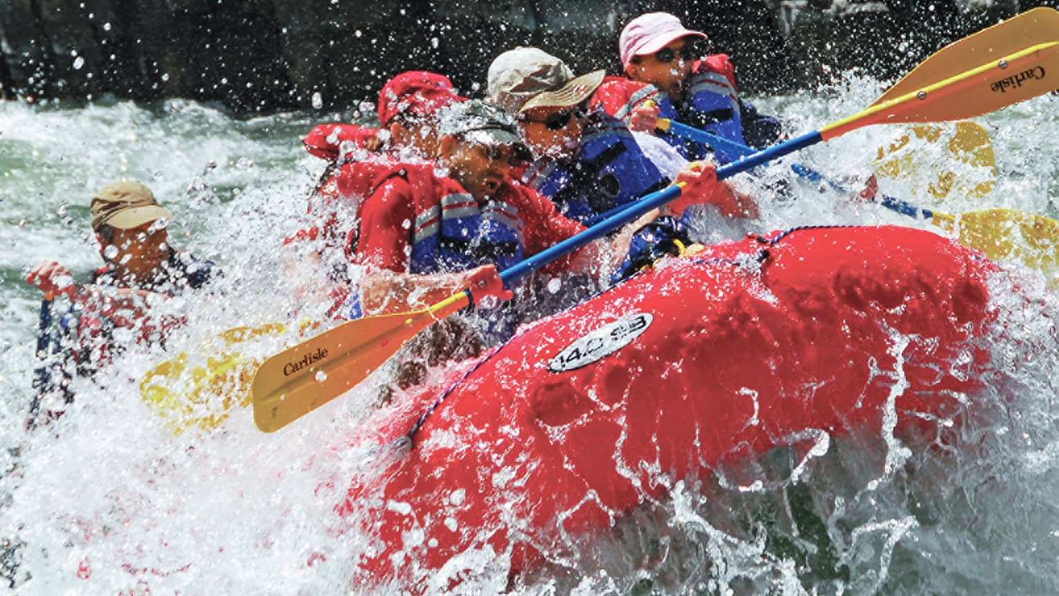 Water sprays around red inflatable raft, people with paddles, life vests white water rafting