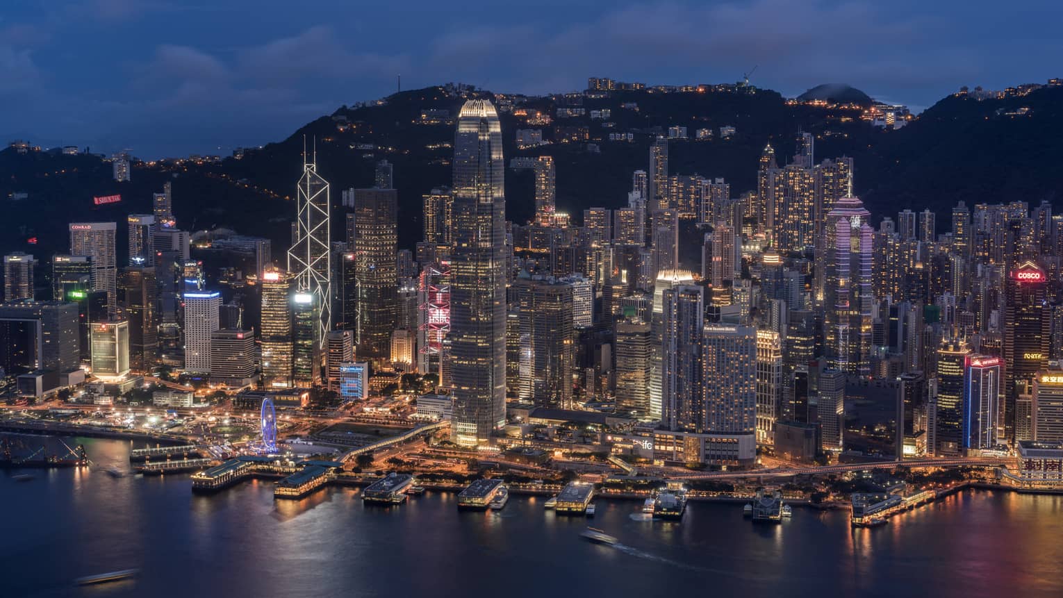 The city skyline at night with buildings of various sizes lit-up by the water’s edge and across the hills in the distance.