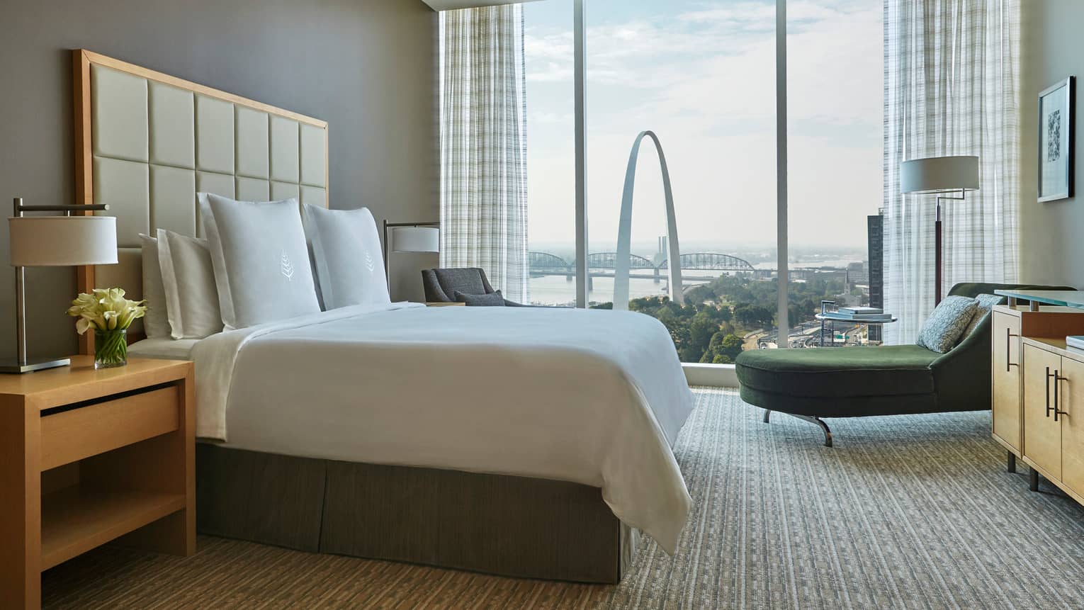 Bed with tall, padded white headboard, green velvet chaise beside floor-to-ceiling window with arch view