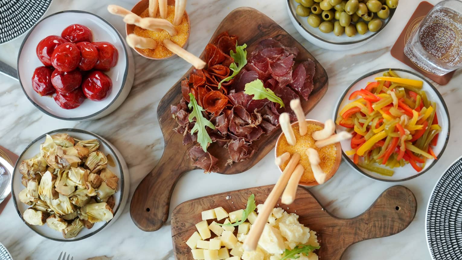 Overhead view of white-and-grey marble table filled with a variety of small plate appetizers, including sliced meats, green olives, sliced cheese, breadsticks and sliced vegetables