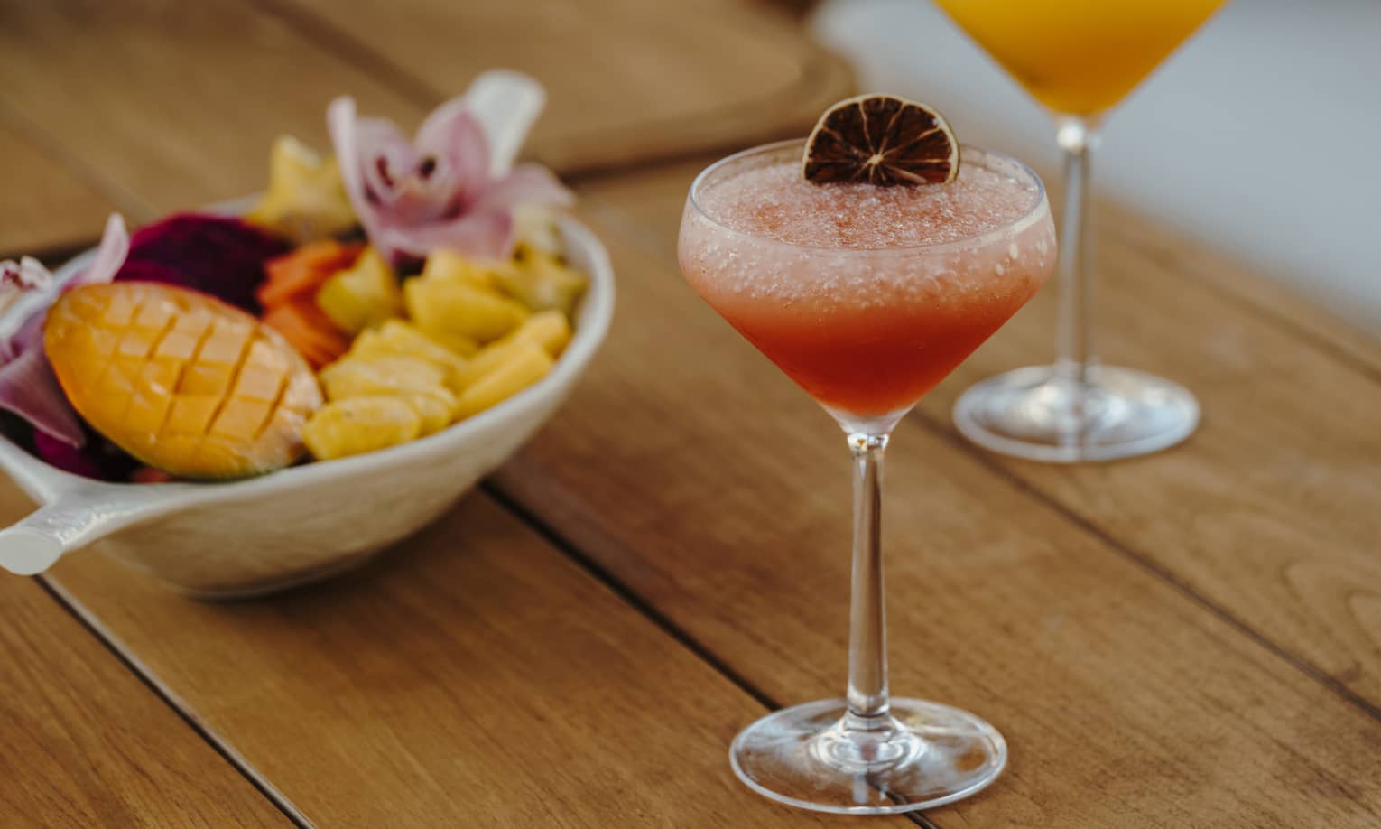 A wood table holds two stemmed glasses of pink and orange drinks with garnishes and a bowl of small-bite food items.