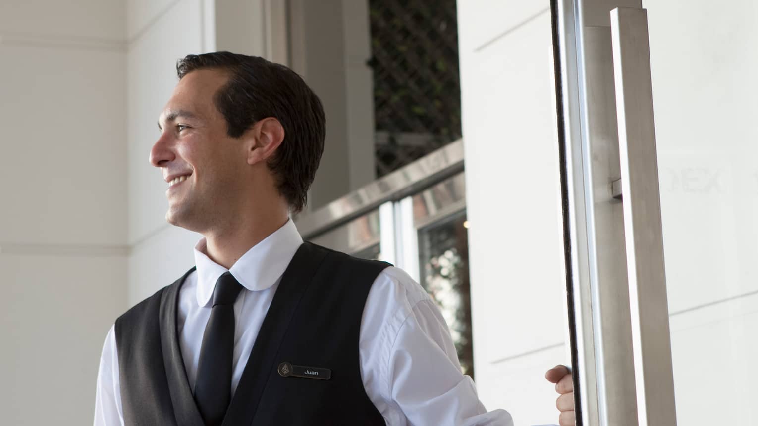 Man in black vest and tie, white shirt smiles and holds door open