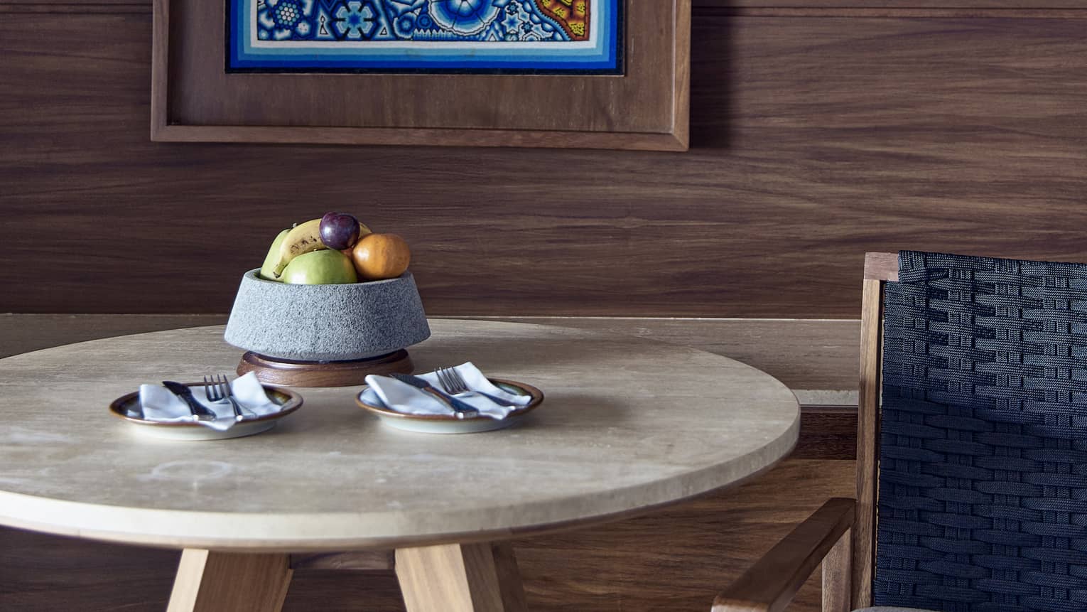 Circular table with bowl of fruit, arm chair, hanging artwork on wooden wall
