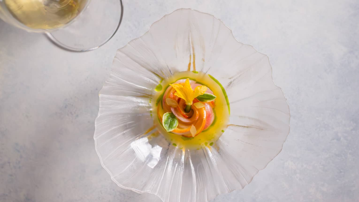 Overhead view of a colourful appetizer garnished with florals in a glass, flower-shaped dish beside a glass of white wine.