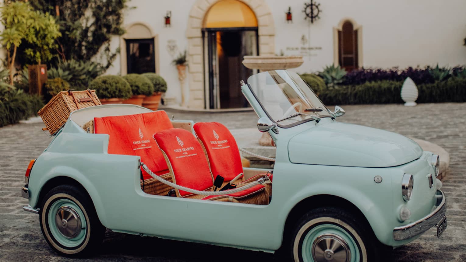 Vintage teal convertible Fiat 500 with bright coral seats parked on a cobblestone driveway in front of San Domenico Palace.  