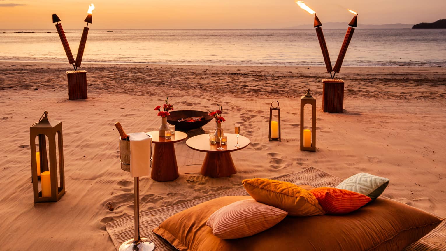Blanket, cushions, small tables, lanterns and tiki torches are set up for a private dinner on a beach at sunset