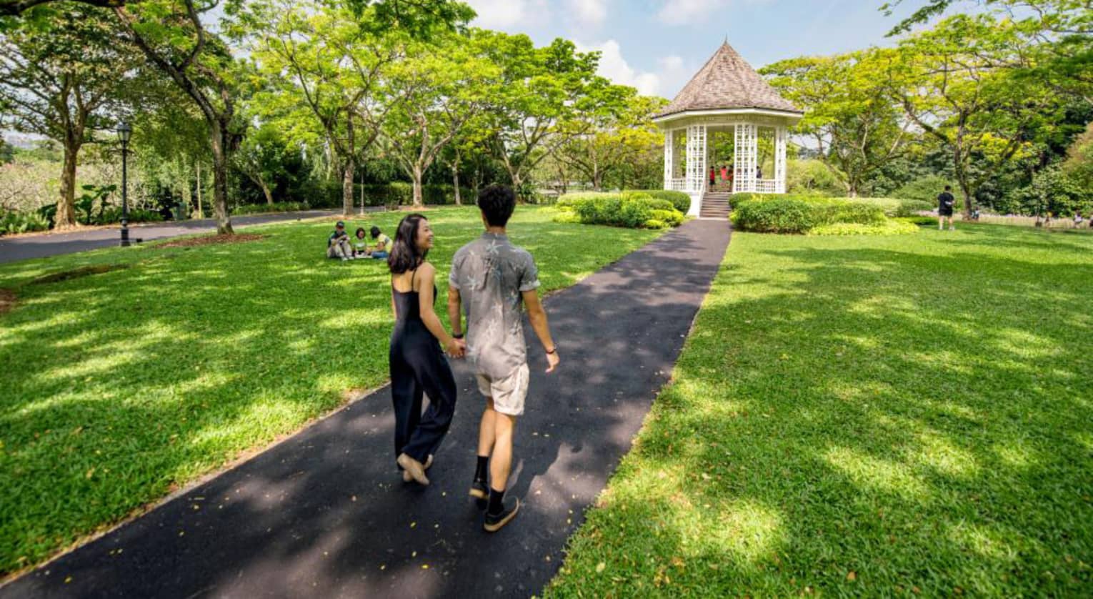 A man and woman hold hands and walk along an asphalt trail through the Singapore Botanic Gardens 