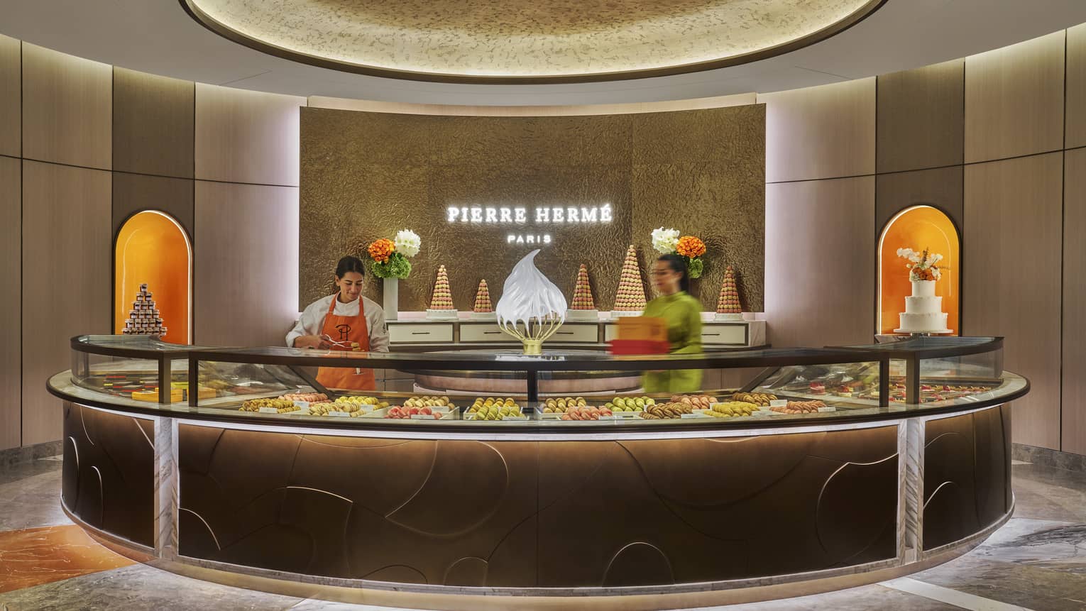 Person wearing white clothing and orange apron stands behind a round, glass-topped pastry counter