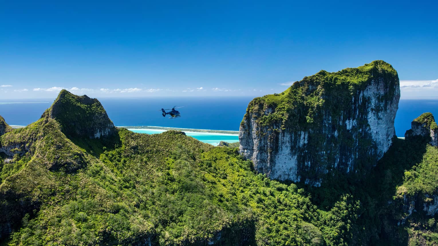 Aerial view of a helicopter flying over forested mountaintops, a turquoise lagoon and the vast open ocean extending beyond.