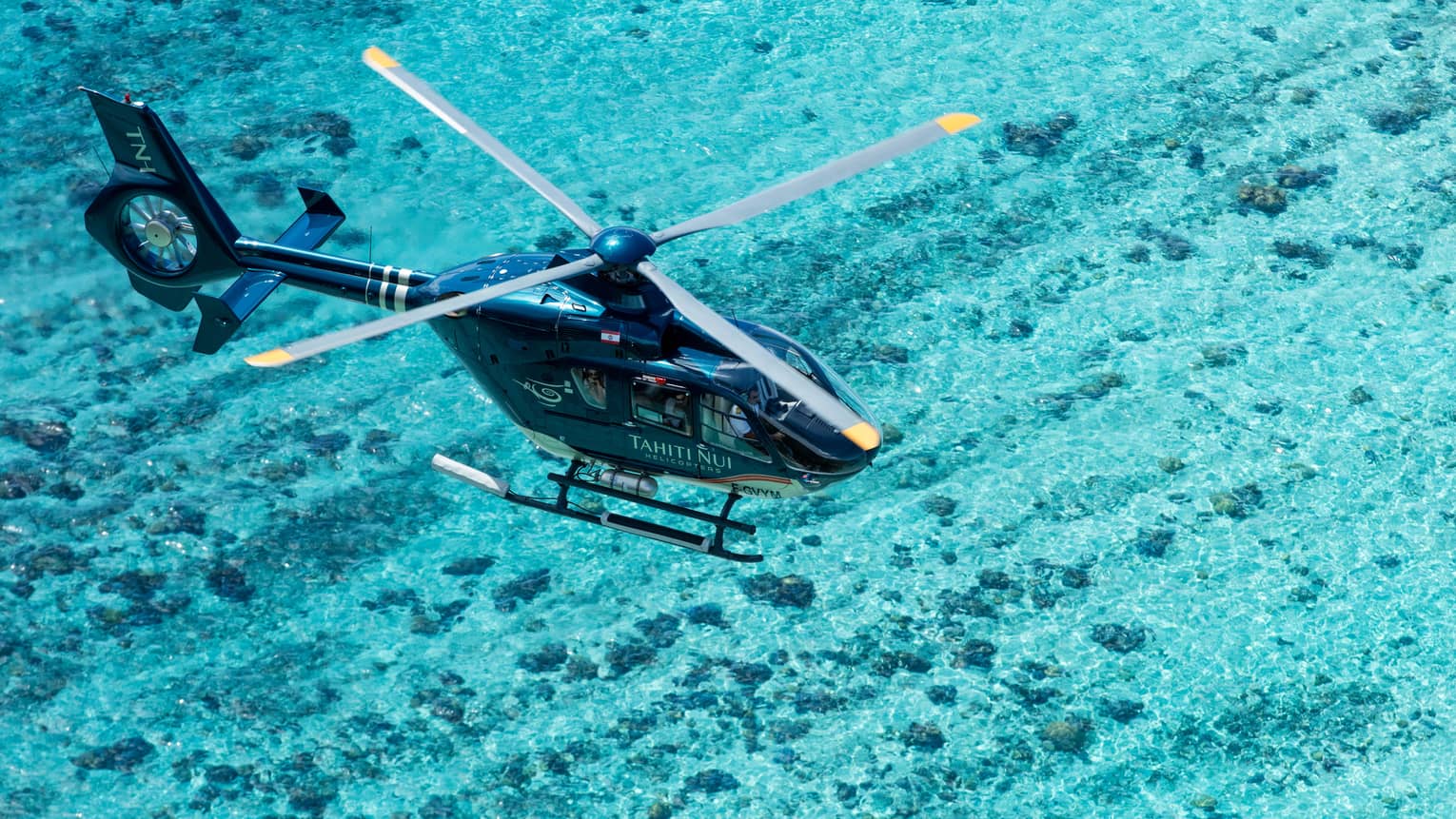 View from above of a helicopter flying over turquoise water so clear the rock ocean floor shows through.