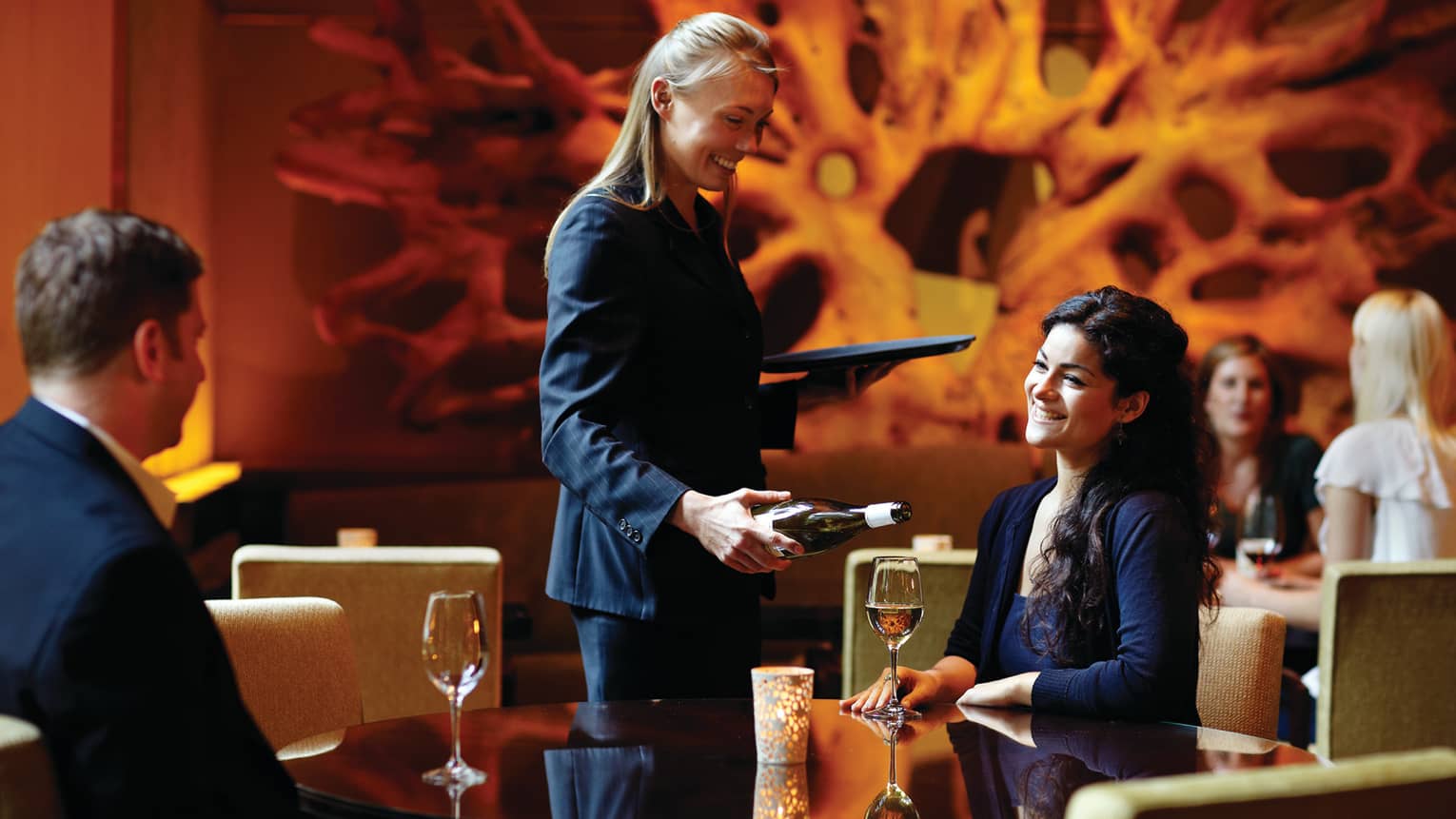 A couple dines at a candlelit table in a restaurant with modern decor, while a server pours wine into one of the glasses on the table