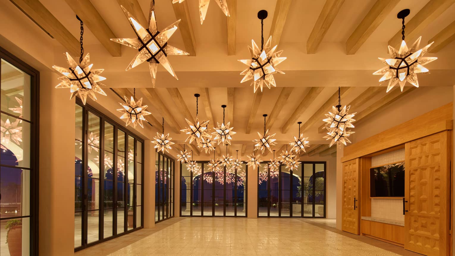 Brightly lit and spacious indoor event space with warm neutral-colour interior, large flat-screen TV with decorative sliding panels, floor-to-ceiling windows, and star-shaped pendant lighting
