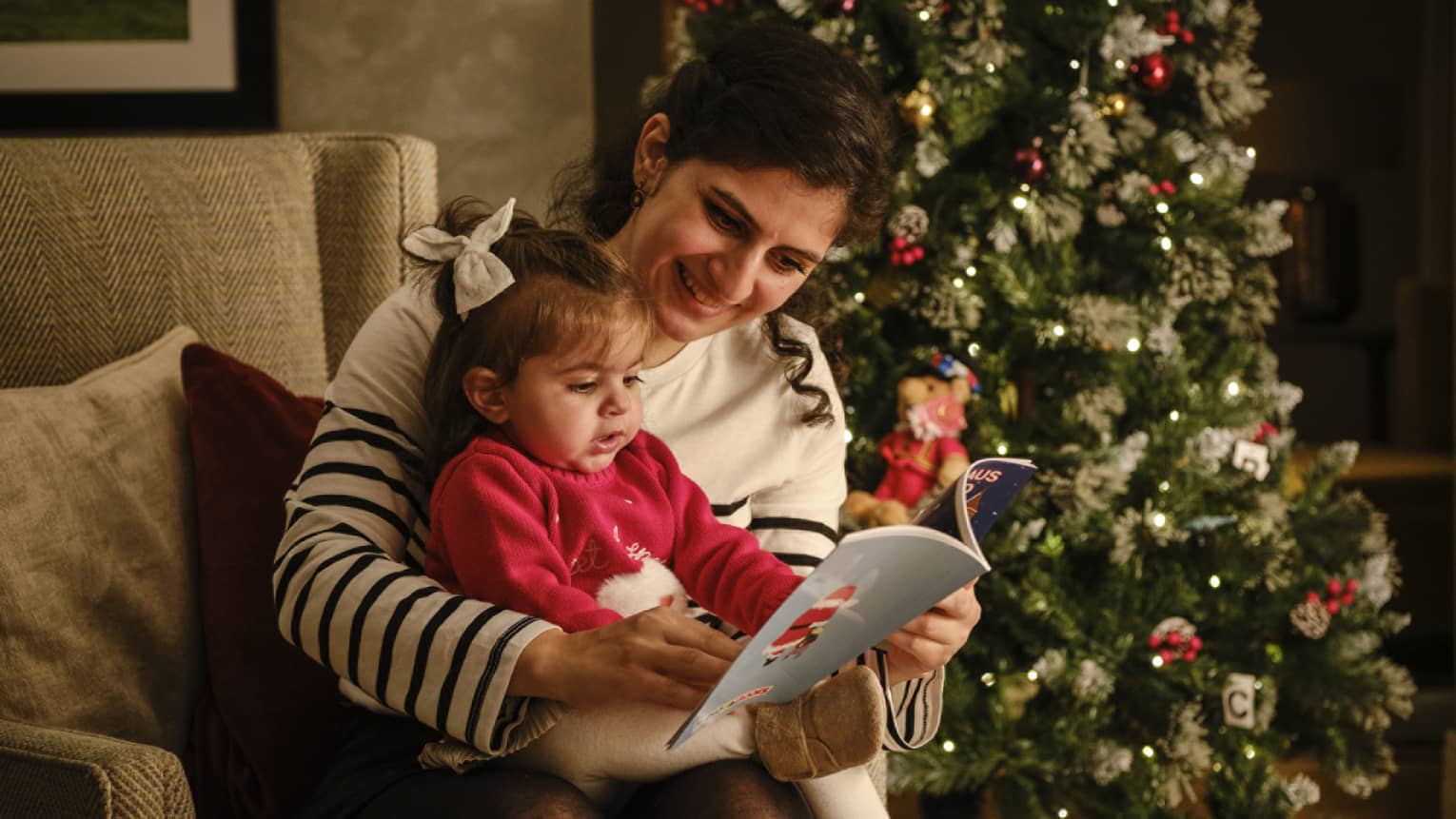 A woman reads a book to a small child while sitting in front of a decorated Christmas tree