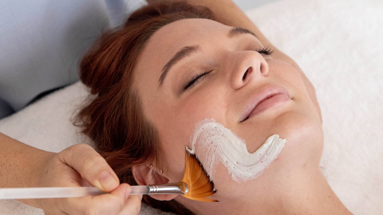 Spa staff paints white cream onto woman's face as she lays with eyes closed