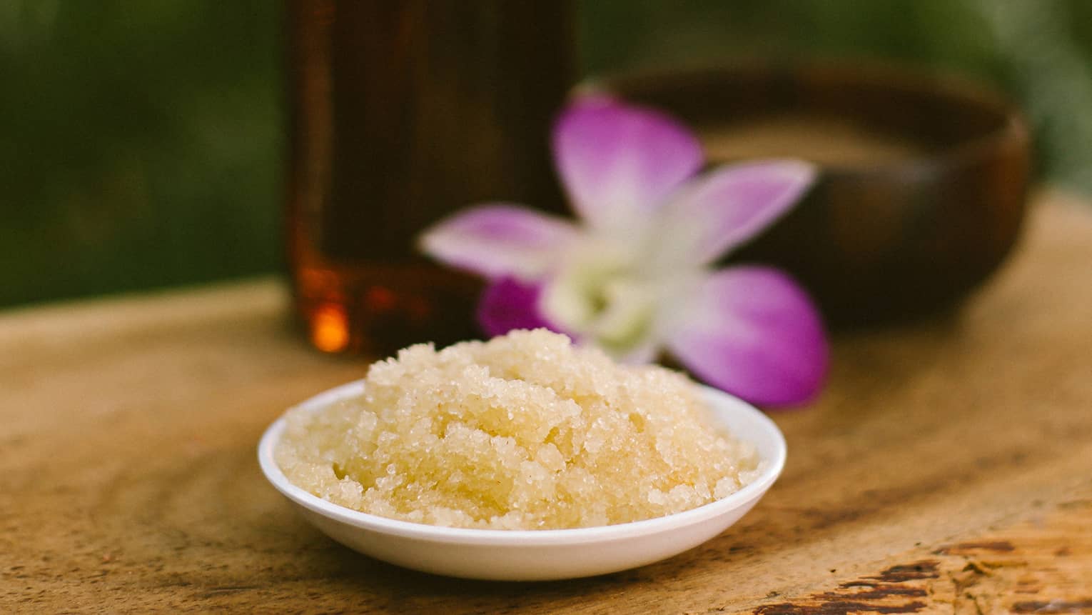 Bowl with salt scrub next to purple tropical flower and glass oil bottle on rustic table