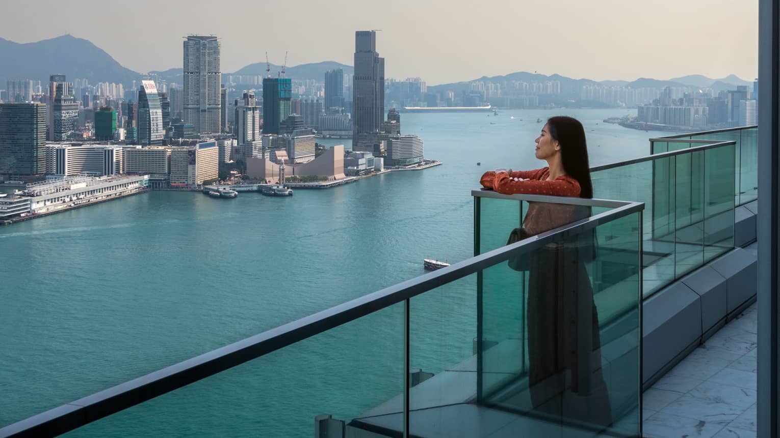 A woman standing on a balcony looking out at water and a city.