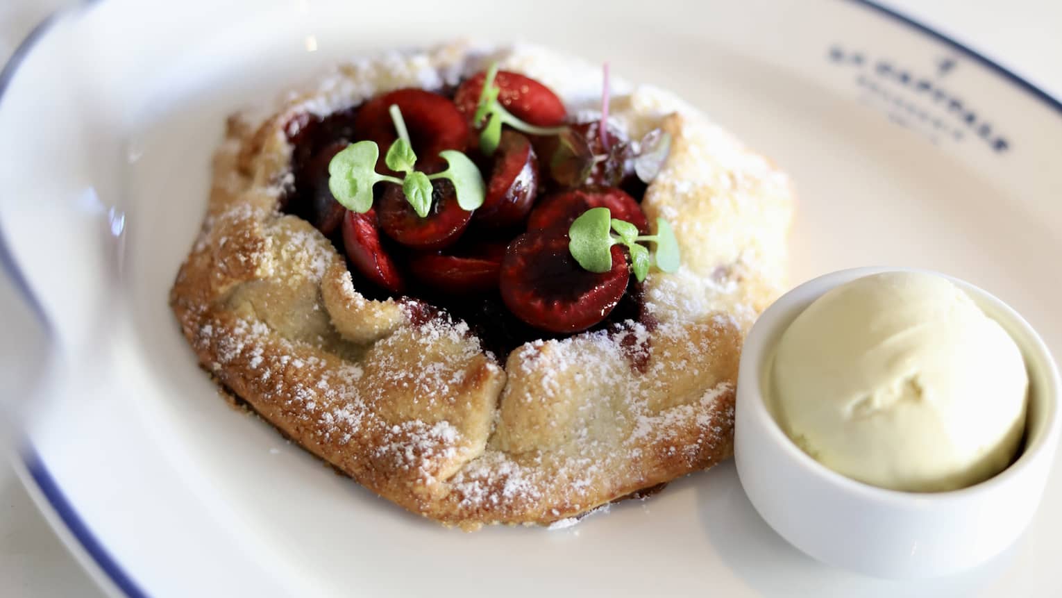Cherry tart garnished with three sprigs of greenery served next to a small scoop of ice cream on a white oval dish