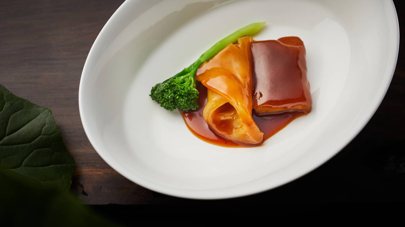 Slice of pork trotter and bright orange fish maw draped in a glossy brown glaze, aside a crisp Chinese broccoli floret. 