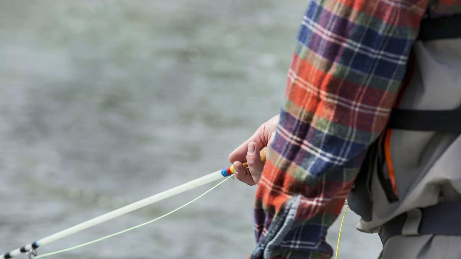 Against a background of rippling silver water, a plaid sleeve partly reveals a hand gently holding a colourful fishing rod.