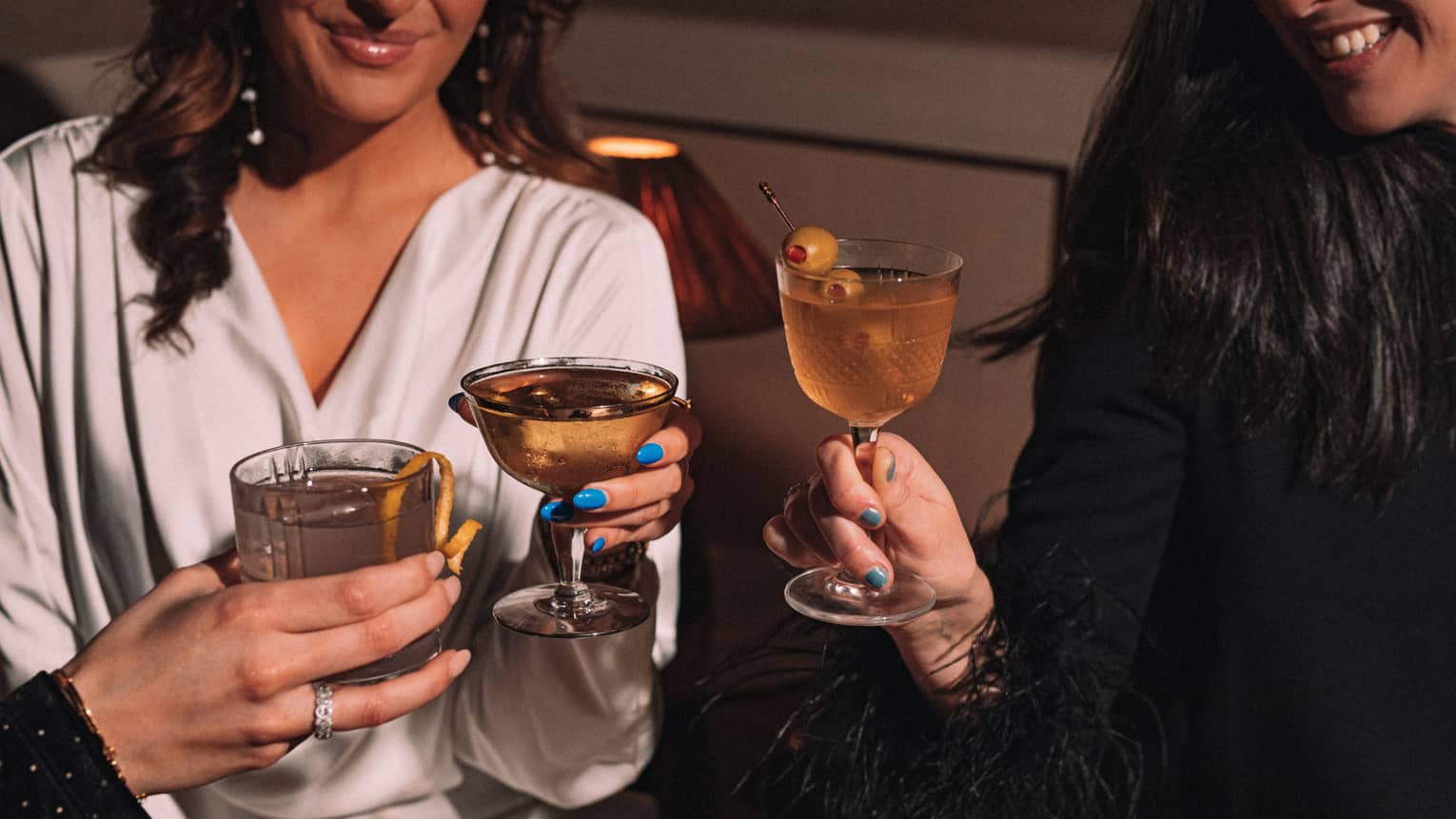 Three smiling guests share a toast with stemmed glasses and tumblers of cocktails with olive and lemon garnish.