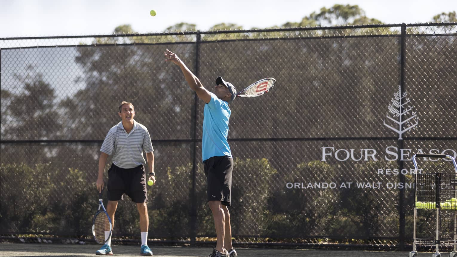 On a tennis court, stray balls are scattered around an instructor demonstrating a serve to a guest who watches intently.
