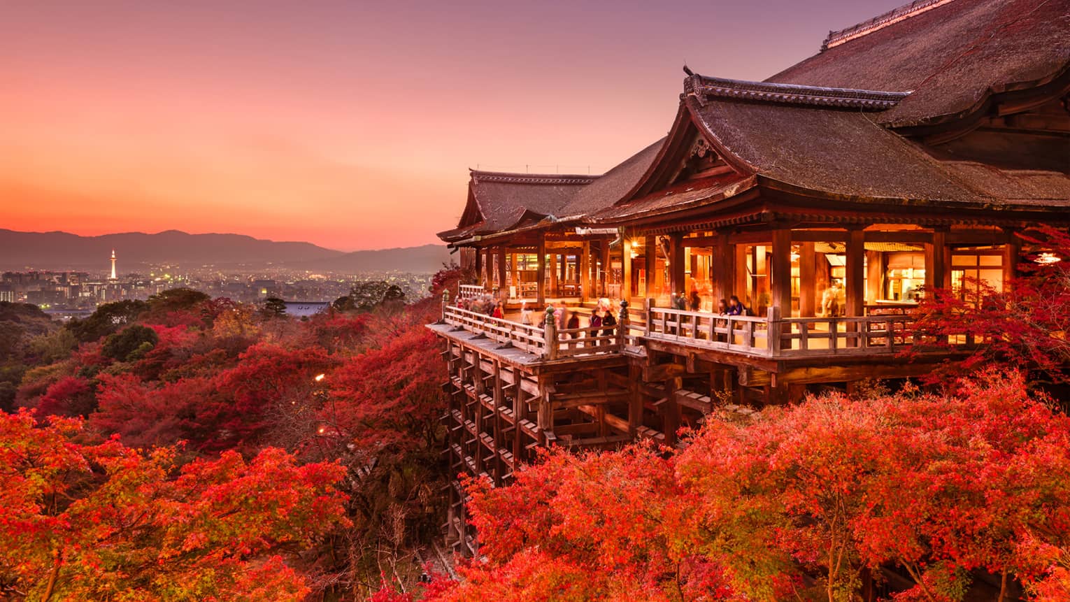 Massive temple on stilts under a pink-hued sky, people on patio admire red-leafed trees, distant city skyline and mountains.