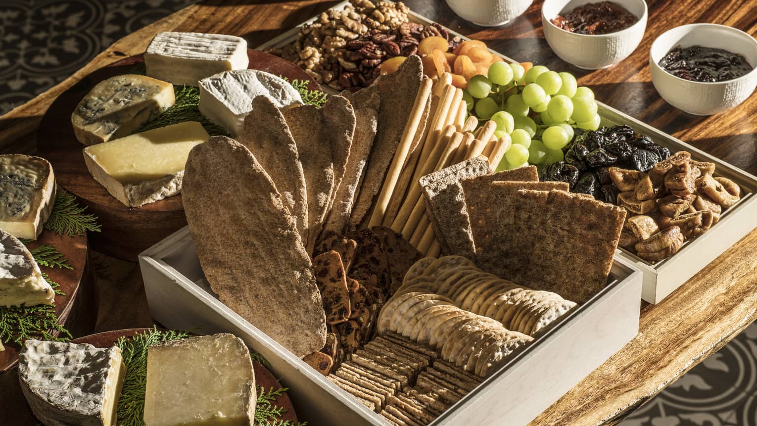 A charcuterie board with crackers and bread, a variety of cheeses, fresh and dried fruit, jams and nuts