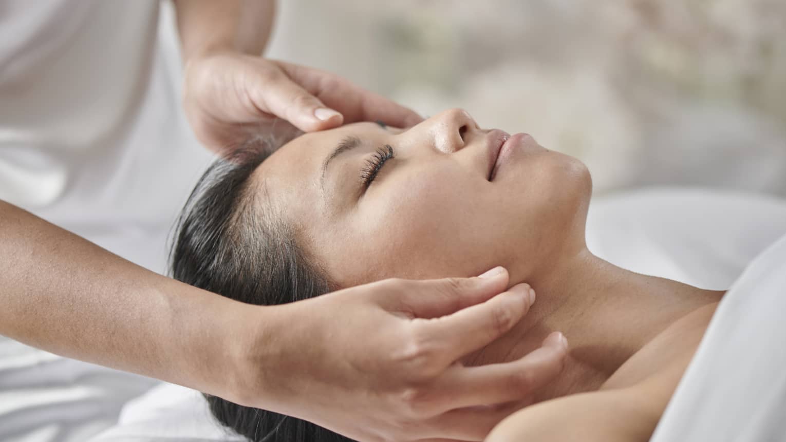 Masseuse massages woman's face as she lies face up on table under white sheet