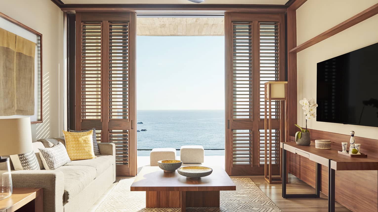 Bright living room with a comfortable grey sofa, wooden furniture and large shutters opening to an ocean view. The room features neutral tones and a modern, airy design.