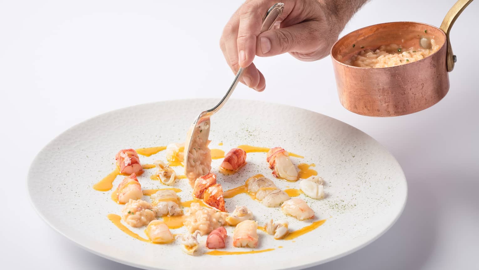 A close up of a hand spooning out a cream sauce and vegetables from a copper ladle onto a porcelain plate.