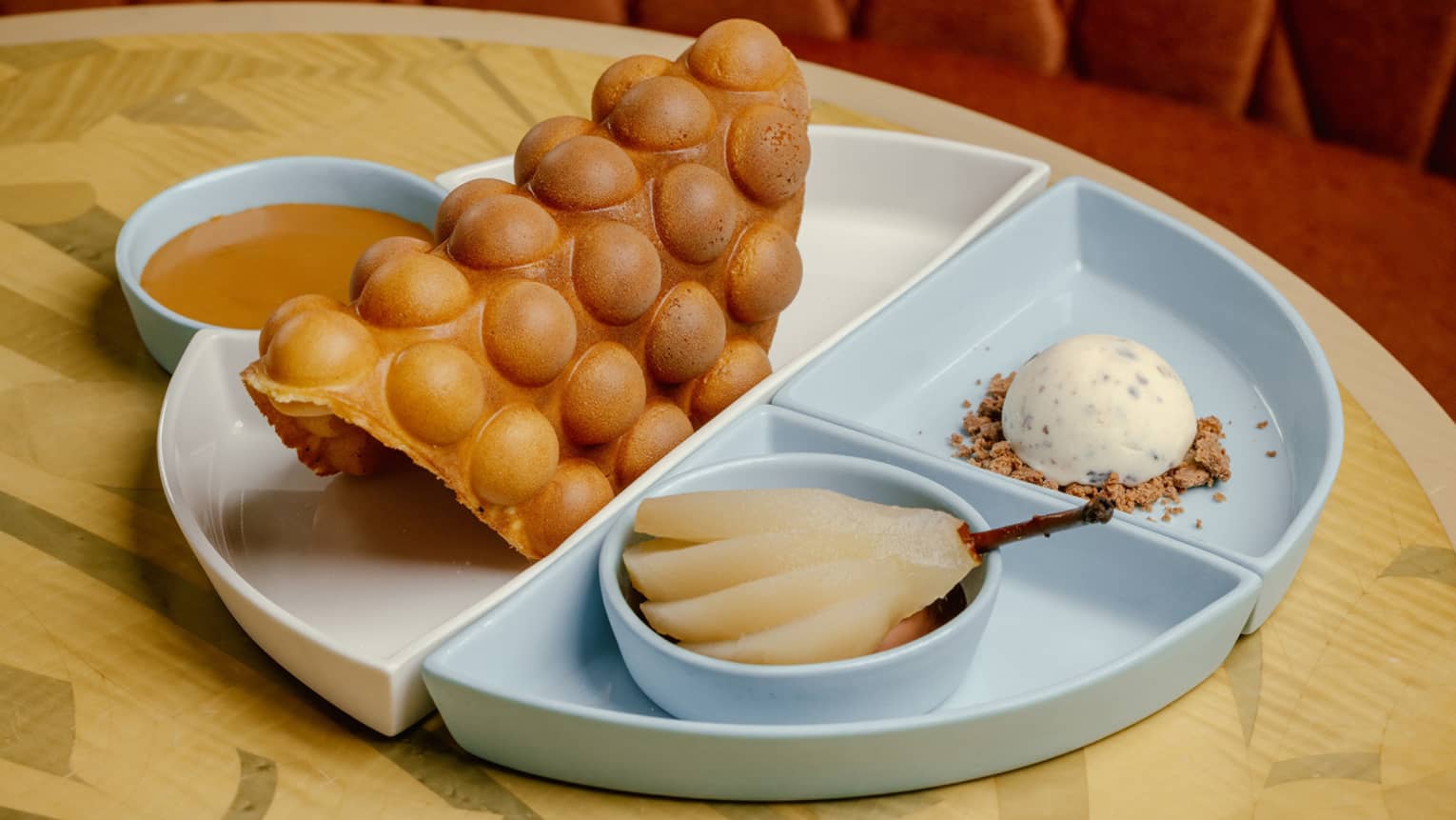 An egg waffle, poached pear, a scoop of ice cream on cookie crumbs and sauce individually and elegantly plated on a table.