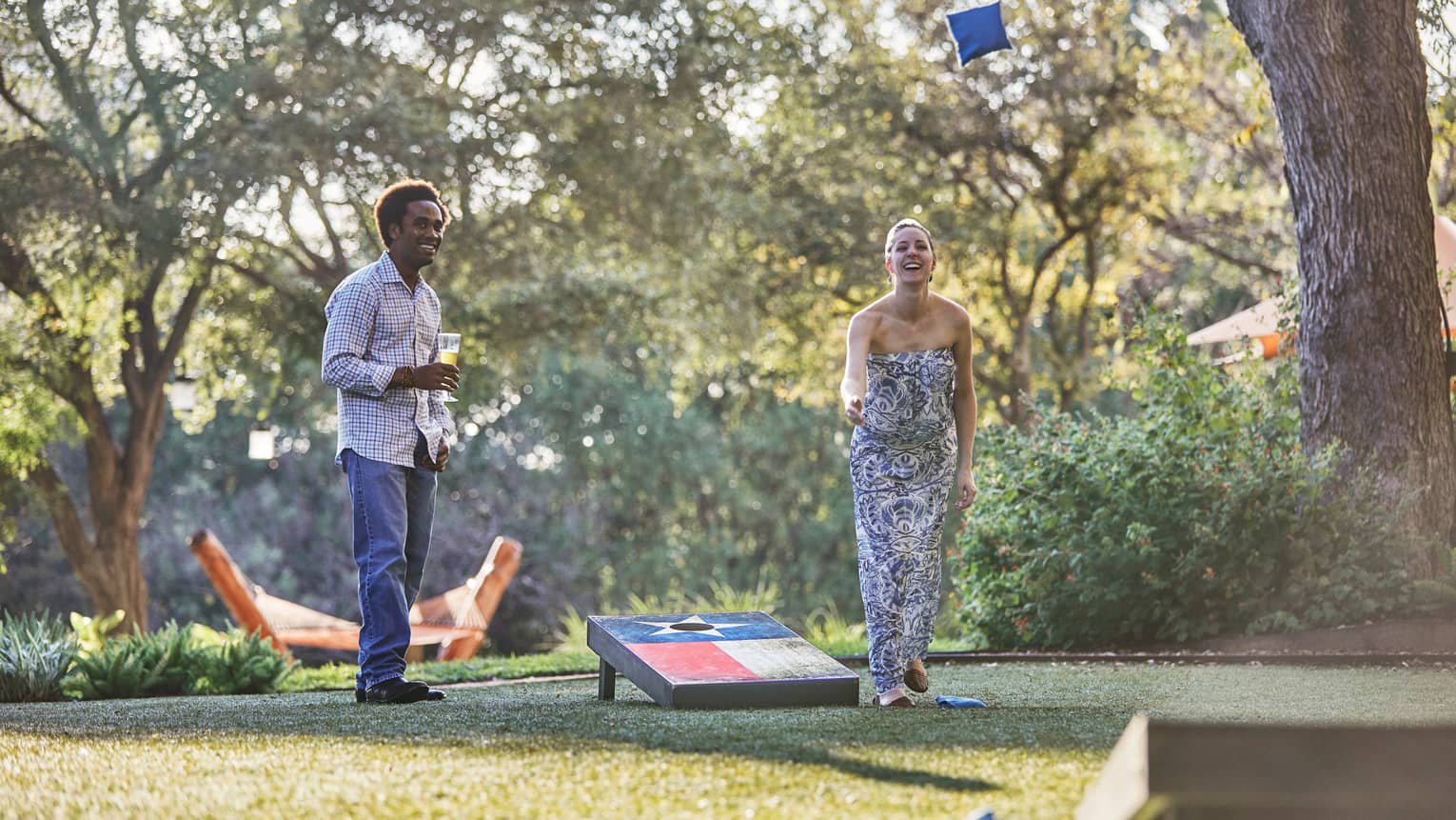 Laughing woman tosses bean bag on lawn, man holding beer watches